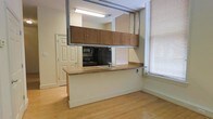 This single-floor space showcases an open-concept kitchen and living area connected by light hardwood flooring that runs throughout, creating a cohesive and easy-to-navigate layout. The kitchen is defined by a peninsula-style counter with a wood-tone laminate surface, offering both workspace and a natural divider between the kitchen and the adjacent living area. Upper cabinetry in warm medium-toned wood is suspended above the peninsula via an exposed industrial-style metal track system, adding a distinctive structural element to the space. Black appliances, including a refrigerator and range, are integrated into the kitchen along with a stainless steel sink. A large window fitted with horizontal blinds allows natural light to filter into the space, brightening the cream-painted walls and pale flooring. Overhead fluorescent lighting supplements natural light. A white paneled door provides access to an adjacent room or entryway. The overall aesthetic blends functional, utilitarian design with modest warmth from the wood-toned finishes, making the space practical and straightforward in its layout.
