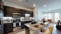 This single-floor residence showcases a thoughtfully designed open-concept layout that seamlessly connects the kitchen, dining, and living areas. The kitchen is a standout feature, outfitted with rich dark espresso cabinetry, light granite countertops, a subtle tile backsplash, and a full suite of stainless steel appliances including a French door refrigerator, range, and built-in microwave. Under-cabinet lighting adds a polished touch, while the central island with granite surface provides additional prep space and casual seating. Wood-look flooring flows throughout the main living areas, complementing the warm, contemporary aesthetic. The dining area comfortably accommodates a small table, and the living room benefits from a ceiling fan, recessed lighting, and large glass doors that invite natural light and provide access to an outdoor balcony with exterior views. The bedroom is generously sized with plush carpeting, multiple windows fitted with blinds and sheer curtains, and access to a private bathroom. The bathroom features a granite-topped vanity with dark cabinetry, a large mirror, and wood-look flooring, with additional access to a walk-in closet. Overall, the space presents a cohesive modern design with quality finishes throughout.