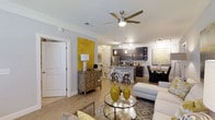 This single-floor residence showcases a thoughtfully designed open-concept layout that effortlessly connects the living, kitchen, and dining areas into one cohesive and functional space. The living area is anchored by a ceiling fan with integrated lighting and is adorned with crown molding, lending a polished, refined feel. A bold yellow accent wall adds a vibrant pop of color that energizes the space and serves as a striking focal point. Light hardwood-style flooring flows throughout, creating a warm and inviting atmosphere. The kitchen is equipped with dark cabinetry, stainless steel appliances including a refrigerator, microwave, and range, as well as a central island with bar seating — ideal for casual dining or entertaining. Pendant lighting and track lighting illuminate the kitchen with purpose and style. The dining area, positioned adjacent to the kitchen, offers ample space for a full dining set and is accented with coordinating artwork. Throughout the space, neutral walls contrast beautifully with carefully chosen décor in yellow and gray tones, creating a cohesive and contemporary aesthetic. The open sightlines between all areas enhance the sense of spaciousness, making this a welcoming and well-appointed single-level home.
