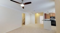This two-story space is designed with an open and functional layout that maximizes usability across both levels. The main living area serves as the heart of the home, featuring plush, light-toned wall-to-wall carpeting that adds warmth and comfort underfoot. Crown molding lines the ceiling, lending a refined architectural detail to the otherwise clean and neutral interior. A ceiling fan with dark wood-toned blades and a built-in light fixture is centrally mounted, providing both airflow and ambient lighting to the space.

The living area flows naturally toward a staircase, providing access to the upper floor. Track lighting in the adjacent hallway area enhances visibility and adds a modern touch. A doorway leads to additional rooms beyond the main living space, suggesting a thoughtful floor plan with clear separation of areas.

The kitchen is partially visible from the living room and features dark, rich wood cabinetry paired with a countertop peninsula or bar-height surface, offering a convenient transition between cooking and living spaces. A sink is positioned near the edge of the counter, adding practicality to the open layout. The overall palette of warm neutrals and light walls creates a bright, welcoming ambiance throughout the main level.
