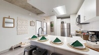 Located at 1401 Curtis St in the heart of Denver, CO, this four-bedroom, three-bathroom unit blends modern design with industrial character across a single floor. The open-concept kitchen is a standout feature, showcasing white upper cabinetry, two-tone lower cabinets in gray, stainless steel appliances including a range, microwave, and refrigerator, and a white quartz peninsula with bar seating — ideal for casual dining and entertaining. A skylight overhead floods the kitchen and living area with natural light, while exposed concrete ceiling beams and columns add a distinctive industrial aesthetic throughout the space. Light wood-style flooring runs continuously through the unit, lending a clean, cohesive feel. The bedrooms are comfortably sized, with the primary bedroom offering ample room for a full furniture arrangement, exposed concrete ceiling accents, and a dedicated workspace area. Additional bedrooms feature large windows that bring in abundant natural light and urban views. The three bathrooms are finished with neutral tile, gray vanities, and shower-tub combinations. A connecting hallway provides efficient flow between rooms. The overall ambiance is modern and airy, combining contemporary finishes with raw architectural details characteristic of urban Denver living.