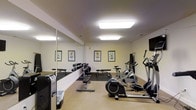 This fitness center offers a practical and well-organized space for residents or guests to maintain their exercise routines. The room is finished with neutral, light-colored walls and carpeted flooring, creating a clean and comfortable environment. Bright recessed fluorescent lighting illuminates the space evenly from above, ensuring excellent visibility throughout.

The cardio section is equipped with multiple machines, including stationary bikes and an elliptical trainer, providing options for low-impact cardiovascular workouts. A wall-mounted flat-screen television is positioned nearby, allowing users to stay entertained during their sessions. A dedicated free weights area features a dumbbell rack with an assortment of weights and accompanying flat benches, suitable for a variety of strength training exercises. Rubber flooring underlays the weight area, offering added safety and floor protection.

A large wall mirror spans one side of the room, enhancing the sense of space while allowing users to monitor their form during workouts. Instructional fitness posters are displayed along the walls, offering exercise guidance. The overall layout is thoughtfully arranged to accommodate multiple users simultaneously with ease.
