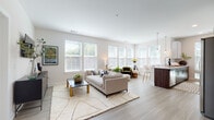 This single-floor residence showcases a thoughtfully designed open-concept layout that seamlessly connects the living, dining, and kitchen areas. The living room is bathed in natural light from an expansive bank of windows dressed with white blinds, highlighting the light-toned hardwood-style flooring that extends throughout the main areas. The space carries a clean, modern aesthetic with neutral walls and generous proportions.

The kitchen is a standout feature, combining white upper cabinetry with rich dark wood lower cabinets and a large peninsula with quartz countertops and bar seating. Globe pendant lights add a contemporary touch, while stainless steel appliances, including a wine refrigerator, complete the well-appointed space. Multiple windows keep the kitchen bright and connected to the outdoors.

The primary bedroom features plush carpeting, a ceiling fan, and large windows that invite natural light. A second bedroom offers an accent wall in deep forest green, creating a cozy, distinct atmosphere. Two bathrooms serve the residence—one featuring a glass-enclosed walk-in shower with warm wood vanity cabinetry, and the other a full soaking tub with elegant grey tile surround. Together, the rooms deliver a cohesive, contemporary living experience.