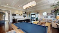 This well-appointed single-floor space functions as an upscale social lounge, combining recreational and leisure areas within an open, cohesive layout. The architecture is defined by prominent coffered ceilings that add depth and a refined character throughout. Rich dark hardwood flooring runs seamlessly across the space, contrasting beautifully with the warm, neutral wall tones.

The recreational zone centers on a full-size pool table with blue felt, positioned beneath a striking rectangular pendant light fixture with organic driftwood-style detailing. Adjacent to this is a generously sized lounge area furnished with a large sectional sofa in neutral tones, accented by blue and yellow throw pillows. A woven rattan coffee table, a bold yellow bench ottoman, and accent chairs complete the seating arrangement, which is anchored by an area rug. A distinctive circular porthole-style window and a curated gallery wall of framed artwork add visual interest to this corner.

The wet bar area features a long white quartz countertop with modern white barstools, dark cabinetry, open display shelving, pendant lighting, a wall-mounted flat-screen TV, and an under-counter refrigerator. Large windows and glass doors throughout invite abundant natural light, and views of lush outdoor greenery enhance the overall bright, airy ambiance of the space.