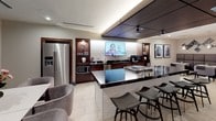 This elegantly designed single-floor space is built for refined entertaining, seamlessly blending a fully equipped kitchen, dining area, wine storage, and lounge into a cohesive, upscale environment. The centerpiece of the space is a chef's kitchen featuring a striking dark granite island with a built-in gas cooktop, illuminated from beneath to highlight its rich veining. Rich mahogany cabinetry lines the back wall, flanking a textured 3D wave-pattern backsplash beneath a wall-mounted flat-screen television. Stainless steel appliances — including a refrigerator, microwave, and professional coffee station — complete the kitchen's functional appeal. A dropped wood-paneled ceiling with integrated track lighting frames the kitchen zone, adding warmth and visual definition. Adjacent to the kitchen, a clean white dining table surrounded by plush grey upholstered chairs provides an inviting setting for formal or casual meals. Two impressive floor-to-ceiling wine display walls, housed in warm wood-framed glass cabinetry, serve as dramatic architectural focal points throughout the space. Beyond, a lounge area with tufted booth-style seating and decorative wall art offers a relaxed retreat. Large porcelain tile flooring in a neutral tone runs throughout, tying the zones together with a polished, contemporary finish.