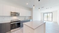 This well-designed single-floor unit offers a cohesive, modern aesthetic throughout, highlighted by light-toned hardwood flooring and a crisp white color palette. The open-plan kitchen and living area serves as the heart of the space, featuring a two-tone cabinetry scheme—upper white cabinets paired with grey lower cabinets—complemented by a white subway tile backsplash and stainless steel appliances, including a range, microwave, dishwasher, and French door refrigerator. A white quartz center island provides additional prep space and casual seating, while modern pendant lighting adds a refined touch overhead. The living area benefits from abundant natural light through a large sliding glass door that opens to a balcony with outdoor views of palm trees and the surrounding streetscape. The bedroom is a well-proportioned, quiet retreat featuring a window with natural views and a dedicated closet. The bathroom presents a clean, contemporary design with a floating grey vanity, white countertop, an undermount sink, and a backlit mirror. An in-unit laundry area is also present. The overall ambiance is bright, clean, and modern throughout.