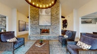 This single-floor amenity facility offers a diverse range of thoughtfully designed spaces for socializing, working, and wellness. The centerpiece is a grand lounge area featuring soaring vaulted ceilings with exposed wood beams, a striking floor-to-ceiling stacked stone fireplace, and a large-format round chandelier with Edison-style bulbs. Comfortable seating arrangements, light hardwood flooring, and curated large-scale artwork create a warm yet refined atmosphere. Adjacent to the lounge, a game room with equally impressive ceiling height houses a pool table and shuffleboard, with a retractable glass garage door that opens the space to the outdoors. The fitness center is comprehensively equipped with Matrix-brand cardio machines including treadmills and cycling equipment, as well as a dedicated strength-training area with cable machines, a Smith machine, and free weights, all set on durable rubberized flooring with mirrored walls. Professional workspaces include a leasing office with wainscoting detail and globe pendant lighting, a glass-enclosed conference room with a chandelier and wall-mounted display, and additional meeting areas with ample natural light. Together, these spaces form a cohesive, upscale community hub designed for convenience and comfort.