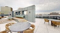 Located atop a prominent building at 1143 Main St in downtown Hartford, Connecticut, this expansive rooftop terrace offers a well-appointed outdoor amenity space with a variety of distinct zones designed for relaxation and social gatherings. The terrace is defined by its modern aesthetic, characterized by sleek gray panel cladding, black metal grid railings, and a mix of large-format porcelain tile and warm wood deck tile flooring throughout.

A generous artificial turf lawn anchors one section of the rooftop, providing a vibrant green contrast to the surrounding hardscape. Adjacent to the turf, a built-in outdoor kitchen features multiple stainless steel grills, prep counters, and ample cabinetry, making it well-suited for outdoor entertaining. Seating areas furnished with woven rattan chairs and round tables are distributed across the terrace, inviting gatherings in multiple settings.

A raised wooden deck platform with stadium-style bench seating adds a unique architectural focal point, while large sliding glass doors lead to an interior rooftop room. Bollard lighting is positioned throughout the space for evening use. The rooftop commands sweeping panoramic views of the Hartford skyline, surrounding neighborhoods, and the verdant landscape beyond, offering an elevated vantage point above the city.