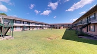 This single-floor residence is part of a well-maintained, two-story courtyard-style complex that opens onto a generous, manicured lawn — a welcoming communal green space framed by the building's U-shaped layout and accessed via exterior staircases. The complex itself features a classic, low-rise architectural design with uniform façades, shuttered windows, and open-air walkways that lend the property a relaxed yet organized character.

Inside, the unit presents a thoughtfully styled interior anchored by polished concrete flooring that runs throughout, adding an industrial warmth to the space. The dining area serves as a visual centerpiece, featuring a striking round marble-topped table paired with woven rattan chairs, all set atop a natural fiber area rug. A large framed abstract artwork adorns the crisp white wall above, contributing to the refined, art-forward aesthetic of the space.

A sliding barn door on a black iron track connects the dining area to an adjacent room, offering both functional flexibility and a design-forward detail. Recessed lighting in the ceiling provides consistent, clean illumination throughout, while visible HVAC vents point to practical, climate-controlled comfort. Neutral white walls throughout keep the interiors bright and versatile, allowing the curated furnishings and artwork to take center stage. The overall ambiance is contemporary and intentional.