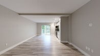 This single-floor unit offers a clean, modern layout designed for efficient and comfortable living. The open-plan living area is finished with light wood-look luxury vinyl plank flooring that runs throughout, complemented by neutral warm-gray and taupe wall tones that create a calm, cohesive aesthetic. Recessed lighting illuminates the space evenly, while large sliding glass doors lead to a private balcony overlooking lush, mature trees, bringing in generous natural light and a refreshing outdoor connection.

The kitchen is integrated seamlessly into the living space and is equipped with stainless steel appliances, including a range and microwave, set against sleek cabinetry that provides practical storage. A hallway connects the main living area to the additional rooms, with a dedicated electrical panel neatly recessed into the wall.

The bathroom is finished in a clean, minimalist style with large-format grey tile flooring, a white vanity with under-cabinet storage, a standard toilet, and a grab bar — suggesting thoughtful, accessible design. Crisp white walls and bright lighting maintain the bright and functional character found throughout the rest of the unit. Overall, this space presents a well-maintained, move-in-ready interior with a neutral palette suited to a variety of personal styles.