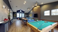 This vibrant recreation lounge is thoughtfully designed to offer a wide range of entertainment options within a single, open-plan space. The room immediately draws the eye upward with its dramatic vaulted ceilings, which lend a sense of grandeur and openness. A diamond-shaped skylight and multiple windows invite natural light to fill the space, complementing the warm wood-look flooring that runs throughout.

At the forefront sits a full-sized pool table with a classic green felt surface, serving as a natural centerpiece for the room. A tall communal bar-height table with seating for several guests provides an ideal spot for casual gatherings or individual work. Toward the back, a cozy lounge area features a blue sectional sofa arranged around a large wall-mounted flat-screen television, set against a striking floor-to-ceiling wood-paneled accent wall in rich warm tones.

The entertainment options continue with arcade game machines tucked along one wall, adding a playful, retro touch to the space. Decorative framed artwork punctuates the deep navy and charcoal-toned walls, contributing to the stylish, modern aesthetic. Recessed lighting and a ceiling fan ensure the space remains comfortable and well-lit throughout the day and evening, making this lounge a versatile hub for socializing and recreation.