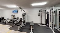 This single-floor fitness room offers a well-rounded selection of exercise equipment arranged across a compact, rectangular space. The room is finished with a combination of rubber mat flooring in the equipment zones and a patterned yellow and grey carpet throughout the remaining floor area, providing both practicality and visual interest. Overhead fluorescent lighting illuminates the entire room, and a textured ceiling adds a subtle design element throughout.

The strength training area is equipped with multiple cable and resistance machines, including a multi-functional cable tower with rope and bar attachments, a leg press/abdominal machine, and an adjustable weight bench. A rack of dumbbells lines one wall, offering a range of free weight options. On the cardio side, the room houses two Matrix treadmills, an elliptical trainer, and a recumbent exercise bike, all positioned on protective rubber mats.

A wall-mounted flat-screen television is positioned for visibility across the equipment area, adding entertainment during workouts. A small storage unit near the entrance holds cleaning supplies and paper towels, supporting hygiene maintenance. A freestanding water cooler near the exit door ensures hydration is easily accessible. The entry door features a distinctive yellow frame, adding a pop of color to the otherwise neutral grey and white interior.