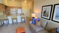 This single-floor space showcases an open-concept design that seamlessly connects the living area and kitchen, creating a cohesive and functional layout. The living area is comfortably appointed with a gray sofa accented by blue and patterned throw pillows, a leather ottoman, and a floor lamp that adds warm ambient lighting. Wall art contributes to the inviting atmosphere, complementing the neutral beige tones found throughout the space. Soft carpeting underfoot enhances the sense of warmth and comfort.

The kitchen is efficiently designed, featuring wooden cabinetry in a warm honey tone, a white refrigerator, and a built-in microwave. A breakfast bar with bar-height seating serves as both a dining option and a natural divider between the kitchen and living areas, offering a practical space for casual meals or entertaining. The countertops feature a neutral stone-like finish that ties the kitchen aesthetic together.

The entryway is visible from the living area, providing a clear sense of the unit's layout and flow. Overall, the space maintains a warm, neutral color palette throughout, creating a welcoming and livable environment.