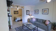 This single-floor space showcases a thoughtful layout that balances openness with defined living zones. The living area serves as the central focus, furnished with a spacious gray sectional sofa paired with a glass-top coffee table resting on a textured area rug. Framed artwork adorns the neutral-toned walls, while a table lamp provides warm ambient lighting, contributing to the room's inviting atmosphere.

A partial dividing wall with decorative transom windows separates the living area from the sleeping space, allowing natural light to pass between the two zones while maintaining a sense of privacy. The sleeping area features a full-sized bed with a floral-patterned comforter, complemented by warm yellow-toned walls that create a cozy, intimate atmosphere. A ceiling fan is installed overhead, serving both spaces efficiently.

The textured ceiling and carpeted flooring run throughout, lending consistency to the overall design. The layout makes efficient use of the available space, with each zone clearly defined yet connected, creating a cohesive and functional living environment suitable for everyday comfort.