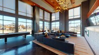 This single-floor community space is designed with a bold contemporary aesthetic that immediately commands attention. Soaring double-height ceilings define the main lounge area, anchored by a striking starburst chandelier with gold-toned arms that serves as a dramatic focal point overhead. An orange accent soffit adds a vibrant pop of color that contrasts beautifully against the dark, polished concrete flooring below.

Floor-to-ceiling windows wrap the perimeter of the space, flooding the interior with generous natural light and providing open views of the surrounding neighborhood. A sliding glass door leads to an outdoor deck with horizontal wood slat railings, extending the living area seamlessly to the exterior.

The main lounge is furnished with expansive sectional sofas in deep charcoal tones, accented with mustard yellow, burnt orange, and navy cushions. A pool table anchors one side of the room, while intimate seating vignettes—featuring curved accent chairs in bold red and yellow hues paired with round bistro tables—are thoughtfully arranged near the windows. Multiple wall-mounted flat-screen televisions are positioned for group viewing, and a built-in fireplace with a white brick surround adds warmth and elegance. A reception desk and a semi-private lounge nook with teal tile accents complete the well-appointed layout.