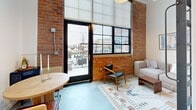 Located at 450 Amsterdam St in Detroit, MI, this efficiently designed one-bedroom, one-bathroom unit makes smart use of its compact footprint across a single floor. The living area is defined by striking exposed brick walls, soaring ceilings with industrial-style glazed transom panels, and a black-framed glass door that opens to an outdoor space, flooding the room with natural light and city views. The living space accommodates a seating area and dining table, anchored by a patterned area rug on polished concrete floors. The galley-style kitchen runs along one wall and is outfitted with light wood cabinetry featuring brass hardware, stainless steel appliances including a full-size refrigerator, a double sink with a sleek faucet, open shelving, and a pendant light — all efficiently arranged to maximize functionality. Exposed ductwork along the ceiling adds an industrial character consistent with the overall aesthetic. The bedroom is a cozy, private retreat with clean white walls accented by wall art, offering a calm and restful atmosphere. The bathroom features a walk-in shower with floor-to-ceiling subway tile, a light wood vanity with a white countertop, and brushed nickel fixtures, completing the unit's cohesive modern-industrial design language.