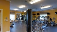 This fitness center offers a comprehensive workout environment designed to accommodate a wide range of exercise needs. The space is finished with warm yellow walls and a neutral rubber flooring, creating an energetic yet functional atmosphere. Overhead, flush-mounted fluorescent lighting and ceiling fans ensure the room remains well-lit and comfortable during workouts.

The cardio section is equipped with multiple treadmills and other aerobic machines, all positioned conveniently near windows that allow natural light to filter in. Flat-screen televisions are mounted on the walls, providing entertainment during cardio sessions. On the strength training side, a multi-station cable machine and a free weight rack with a full mirror backdrop offer users a dedicated area for resistance and muscle-building exercises.

Large windows along the perimeter bring in ample daylight and offer views to the outside, contributing to an open and airy feel. The layout is thoughtfully organized to allow users to move freely between equipment without congestion. Whether focusing on cardiovascular endurance, strength, or flexibility, this single-floor fitness center is designed to support a well-rounded fitness routine in a clean and inviting setting.