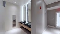 This well-appointed restroom showcases a refined, contemporary design that prioritizes both functionality and visual appeal. The vanity area serves as the focal point, featuring a floating wood-toned counter finished in a warm natural tone and set against a dark surface that provides striking contrast. Two square vessel sinks — one in dark stone and one in translucent glass — are paired with polished chrome faucets, lending an upscale, boutique-style character to the space.

Tall, backlit mirrors extend nearly floor to ceiling, flanking a decorative vertical mosaic tile column that adds texture and visual interest between the reflective panels. The backlighting creates a soft, even illumination that enhances the overall brightness of the room. Recessed ceiling lights complement this lighting scheme, maintaining a clean, uncluttered overhead appearance.

The flooring consists of large-format light-toned tiles, contributing to the space's open and airy feel. Neutral warm-gray walls keep the palette cohesive and understated. A fire alarm panel is mounted discreetly on the wall, indicating a commercial or institutional setting. The overall ambiance is polished and modern, reflecting a high standard of interior design.