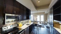 This single-floor residence showcases a thoughtfully designed open-concept layout that seamlessly connects the kitchen, dining, and living areas. The kitchen is a standout feature, outfitted with rich, dark espresso cabinetry that extends to the ceiling, offering generous storage. Light granite countertops provide a striking contrast, while a mosaic tile backsplash adds texture and visual interest. Stainless steel appliances, including a built-in microwave, electric range, and dishwasher, complete the modern culinary space. A double undermount sink and under-cabinet lighting further enhance functionality. The kitchen flows naturally into the dining area, where a compact table with bar-height seating sits beneath elegant pendant lighting. The living area beyond benefits from an abundance of natural light streaming through large French doors, creating a bright and welcoming atmosphere. Dark hardwood flooring runs throughout, anchoring the space with warmth and sophistication. A tray ceiling detail above the kitchen adds architectural depth and dimension. The overall ambiance is contemporary and polished, with a cohesive neutral color palette accented by bold dark tones — ideal for both everyday living and entertaining.
