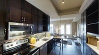 This single-floor residence showcases a thoughtfully designed open-concept layout that seamlessly connects the kitchen, dining, and living areas. The kitchen is a standout feature, outfitted with rich, dark espresso cabinetry that extends to the ceiling, offering generous storage. Light granite countertops provide a striking contrast, while a mosaic tile backsplash adds texture and visual interest. Stainless steel appliances, including a built-in microwave, electric range, and dishwasher, complete the modern culinary space. A double undermount sink and under-cabinet lighting further enhance functionality. The kitchen flows naturally into the dining area, where a compact table with bar-height seating sits beneath elegant pendant lighting. The living area beyond benefits from an abundance of natural light streaming through large French doors, creating a bright and welcoming atmosphere. Dark hardwood flooring runs throughout, anchoring the space with warmth and sophistication. A tray ceiling detail above the kitchen adds architectural depth and dimension. The overall ambiance is contemporary and polished, with a cohesive neutral color palette accented by bold dark tones — ideal for both everyday living and entertaining.