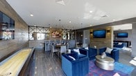 The clubroom at 665–711 Commerce Dr in Fairfield, NY, is a thoughtfully designed communal space that blends contemporary style with practical amenities. The single-floor layout is organized into distinct zones, allowing for both social gatherings and relaxed lounging. A prominent lounge area anchors one side of the room, furnished with plush royal blue velvet seating and a sleek round coffee table, positioned atop a richly colored area rug that adds visual warmth to the space. A wall-mounted flat-screen TV is positioned nearby, making it an ideal spot for casual viewing or entertainment. Toward the back, a bar area features high-top seating with modern bar stools arranged along counter-height tables, complemented by pendant lighting with gold-toned fixtures that lend an upscale ambiance. Wood-paneled accent walls add texture and warmth, contrasting with the cool gray tones of the large-format tile flooring. Geometric metal detailing on the walls and decorative mirrors enhance the room's modern character. Recessed lighting throughout ensures consistent illumination, while the open layout promotes easy flow between seating zones, making this clubroom a versatile and inviting gathering space.