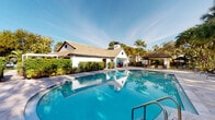 This single-story property is defined by its standout outdoor amenity: a generously sized swimming pool with clear, vivid blue water that serves as the centerpiece of the exterior. The pool area is well-appointed with a broad surrounding deck, providing ample space for lounging and recreation. A classic white clubhouse structure with a pitched, tile roof and a distinctive circular window sits directly adjacent to the pool, offering a clean architectural contrast against the lush tropical landscaping. A covered gazebo nearby provides a shaded outdoor gathering space, ideal for relaxing away from direct sun. Mature palm trees and dense greenery form a natural perimeter around the pool area, enhancing the sense of privacy and creating a distinctly tropical ambiance. The bright Florida sky and abundant natural sunlight contribute to the open, resort-like atmosphere of the outdoor space. The remaining interior images do not provide sufficient detail to describe the indoor areas of the property. Overall, the outdoor environment conveys a well-maintained, amenity-rich setting with a warm, sun-drenched character consistent with a tropical residential community.