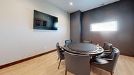 Located at 1415 County Road B W in Roseville, MN, this dedicated card room is a purpose-built space designed for card games and social gatherings. The room features light hardwood flooring throughout, complemented by a two-tone wall design that pairs light gray upper walls with a darker charcoal accent, lending the space a modern and polished aesthetic. Recessed ceiling lighting provides even, comfortable illumination across the room.

The centerpiece of the space is a high-quality round poker table surrounded by upholstered chairs, accommodating multiple players in a comfortable and stylish setting. A wall-mounted flat-screen television is positioned for easy viewing, adding a multimedia element ideal for entertainment during gameplay.

The walls are adorned with a curated collection of framed sports venue artwork, including prints of notable Minnesota stadiums such as U.S. Bank Stadium, Target Field, and Xcel Energy Center, giving the room a distinctly local character. A small horizontal window allows a modest amount of natural light to enter, while a solid wood interior door with a door closer provides privacy. Safety features, including a fire alarm and security camera, are also present. The overall ambiance is intimate, modern, and purpose-driven — well-suited for an enjoyable card game experience.
