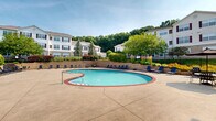 Located at 1660 Hampton Knoll Dr in Akron, Ohio, this outdoor amenity space centers around a generously sized kidney-shaped swimming pool with a classic brick coping border and clear turquoise water. The expansive concrete sundeck surrounds the pool on all sides, providing ample room for lounging and socializing. Numerous blue lounge chairs and patio seating arrangements are thoughtfully positioned throughout the deck, offering residents plenty of options for relaxation and outdoor dining.

Adjacent to the pool, a well-maintained single-story clubhouse building features large windows, white exterior siding, and a charming chimney, providing a functional and inviting gathering space. Umbrella-shaded tables near the clubhouse offer additional outdoor seating in a shaded setting.

The pool area is enclosed by an iron fence and framed by raised stone retaining walls with ornamental grasses and seasonal plantings, adding both privacy and visual appeal. Mature trees and lush landscaping surround the perimeter, creating a green, park-like atmosphere. In the background, multi-story residential buildings with distinctive burgundy shutters and cream-colored exteriors are visible, set against a backdrop of wooded hillside. The overall setting is bright, open, and well-appointed, making it an ideal outdoor retreat for residents.