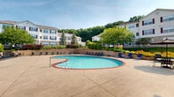 Located at 1660 Hampton Knoll Dr in Akron, Ohio, this outdoor amenity space centers around a generously sized kidney-shaped swimming pool with a classic brick coping border and clear turquoise water. The expansive concrete sundeck surrounds the pool on all sides, providing ample room for lounging and socializing. Numerous blue lounge chairs and patio seating arrangements are thoughtfully positioned throughout the deck, offering residents plenty of options for relaxation and outdoor dining.

Adjacent to the pool, a well-maintained single-story clubhouse building features large windows, white exterior siding, and a charming chimney, providing a functional and inviting gathering space. Umbrella-shaded tables near the clubhouse offer additional outdoor seating in a shaded setting.

The pool area is enclosed by an iron fence and framed by raised stone retaining walls with ornamental grasses and seasonal plantings, adding both privacy and visual appeal. Mature trees and lush landscaping surround the perimeter, creating a green, park-like atmosphere. In the background, multi-story residential buildings with distinctive burgundy shutters and cream-colored exteriors are visible, set against a backdrop of wooded hillside. The overall setting is bright, open, and well-appointed, making it an ideal outdoor retreat for residents.