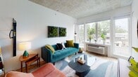 This single-floor unit showcases a contemporary design characterized by striking exposed concrete ceilings that lend an industrial-chic aesthetic throughout the space. Light wood flooring runs continuously across the main areas, creating warmth that contrasts effectively with the raw ceiling texture.

The kitchen is thoughtfully designed with rich dark wood cabinetry, both upper and lower, complemented by clean white countertops and a hexagonal tile backsplash. A full suite of stainless steel appliances includes a gas range with oven, over-the-range microwave, dishwasher, and full-size refrigerator. Open shelving provides additional display and storage space.

The living area is bright and welcoming, with large corner windows that frame urban views and fill the room with natural light. The space easily accommodates a seating arrangement and media console.

The bedroom benefits from expansive floor-to-ceiling windows offering outdoor views, and includes a dedicated walk-in closet with built-in rod and shelving. The bathroom features wood-toned cabinetry, a combined tub and shower with tile surround, and wood-look flooring, maintaining the unit's cohesive modern palette throughout.