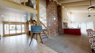 Located at 1401 E Girard Pl in Englewood, Colorado, this two-story facility is defined by its striking architectural character and versatile amenities. The grand entry lobby immediately commands attention with soaring ceilings, exposed white-painted beam work, and a prominent full-height brick column that anchors the open floor plan. Tile flooring transitions seamlessly to patterned carpet as the space flows from the entry toward a central staircase connecting both levels.

The main floor features a well-appointed kitchen with rich wood cabinetry, granite countertops, a center island, and full appliances — ideal for community gatherings. Adjacent to the kitchen, a comfortable lounge area offers vaulted ceilings with exposed beams, plush leather seating, and wall-mounted display screens for entertainment or presentations.

A dedicated fitness room is equipped with free weights, a cable machine, a treadmill, wall mirrors, and high windows that bring in natural light. The business center provides computer workstations with ergonomic seating in a private, glass-partitioned area. Upper-level office spaces overlook the main floor through glass walls, enhancing the open, connected feel. A secondary workspace with direct access to an outdoor deck rounds out the upper level, offering views of the surrounding landscape.