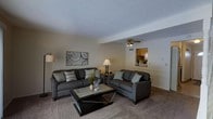 This three-floor property offers a comfortable and functional layout designed to maximize both space and livability. The main living area is a welcoming space anchored by plush, dark gray upholstered sofas arranged around a rustic wood-and-metal coffee table, creating an inviting focal point for relaxation and entertaining. Neutral beige carpeting and warm-toned walls contribute to a calm, cohesive ambiance throughout the room. A ceiling fan overhead provides both practical airflow and a subtle design element. Decorative touches, including framed abstract wall art and tasteful accent pieces, add personality to the space. A distinctive architectural pass-through opening connects the living area to the kitchen, where warm wood cabinetry is visible, enhancing the open and communicative feel between the two spaces. The layout across three floors suggests a well-organized separation of living, sleeping, and utility spaces, offering residents both communal areas and private retreats. Overall, the property presents a balanced blend of practicality and comfort, with consistent neutral finishes that lend themselves well to personalization.
