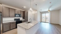 Located in Denton, TX, this efficiently designed 1-bedroom, 1-bathroom residence offers a thoughtful single-floor layout that maximizes every square foot. The open-concept living area seamlessly connects the kitchen, dining, and living spaces, creating an airy and functional atmosphere. The kitchen is a standout feature, showcasing rich dark-toned cabinetry, granite-look countertops, black appliances including a refrigerator, range, and microwave, and a central island with a built-in sink illuminated by stylish pendant lighting. Light wood-look flooring flows throughout the main living areas, enhancing the bright, clean aesthetic. A sliding glass door in the living area allows natural light to pour in while providing outdoor views. The bedroom offers soft carpeting underfoot, a ceiling fan, and a window with neighborhood views. The full bathroom features a dual-sink vanity with granite countertops, wood-look flooring, and a tiled tub-shower combination. Direct access leads to a generously sized walk-in closet outfitted with multi-tiered wire shelving for ample storage. Additional utility and laundry spaces round out this well-planned residence, making it a practical and comfortable living environment.
