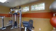 This single-floor fitness center is designed to provide a functional and motivating workout environment. The space is defined by its bold, striped accent walls — featuring warm golden-yellow and deep terracotta-orange horizontal bands — which lend the room an energetic and dynamic character. A row of clerestory windows runs along the upper portion of the wall, allowing natural light to filter in while framing views of surrounding trees, creating a refreshing connection to the outdoors.

The facility is outfitted with a selection of professional-grade exercise equipment, including a multi-function strength training cable machine and a Cybex stationary cycle, indicating a well-rounded approach to both resistance and cardiovascular training. The flooring features a durable, low-pile carpet suited for a high-traffic fitness environment.

The overall layout is thoughtfully organized to maximize usable floor space while maintaining an open, accessible feel. The neutral ceiling complements the vibrant wall colors, keeping the visual energy grounded. The combination of natural light, bold design choices, and quality equipment makes this fitness center an inviting and practical space for a comprehensive workout routine.