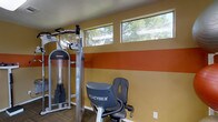 This single-floor fitness center is designed to provide a functional and motivating workout environment. The space is defined by its bold, striped accent walls — featuring warm golden-yellow and deep terracotta-orange horizontal bands — which lend the room an energetic and dynamic character. A row of clerestory windows runs along the upper portion of the wall, allowing natural light to filter in while framing views of surrounding trees, creating a refreshing connection to the outdoors.

The facility is outfitted with a selection of professional-grade exercise equipment, including a multi-function strength training cable machine and a Cybex stationary cycle, indicating a well-rounded approach to both resistance and cardiovascular training. The flooring features a durable, low-pile carpet suited for a high-traffic fitness environment.

The overall layout is thoughtfully organized to maximize usable floor space while maintaining an open, accessible feel. The neutral ceiling complements the vibrant wall colors, keeping the visual energy grounded. The combination of natural light, bold design choices, and quality equipment makes this fitness center an inviting and practical space for a comprehensive workout routine.