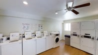 This shared laundry facility is designed to accommodate multiple users simultaneously, offering a well-organized and functional space. A row of top-loading and front-loading washing machines lines one wall, complemented by stacked dryer units positioned along an adjacent wall, providing ample capacity for residents. The room features hardwood-style flooring that is both durable and easy to maintain, contributing to a clean and practical aesthetic. A ceiling fan with an integrated light fixture occupies the center of the room, promoting air circulation and keeping the space comfortable during use. Recessed lighting adds further brightness throughout. A window along one wall allows natural light to filter into the room, enhancing the overall ambiance. Instructional posters are mounted on the wall, offering clear guidance on washing and drying procedures to ensure proper machine use. The walls are finished in a neutral, light tone, giving the space an open and clean feel. The layout is thoughtfully arranged to allow easy movement between machines, making the laundry process efficient and straightforward for all users.