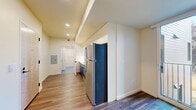 This thoughtfully designed single-floor unit makes efficient use of its compact footprint, offering a clean, modern aesthetic throughout. The kitchen is a standout feature, showcasing bold navy blue lower cabinetry paired with crisp white upper cabinets that extend to the ceiling for maximum storage. A full suite of stainless steel appliances — including a refrigerator, electric range, over-the-range microwave, and dishwasher — provides full functionality. A single-basin sink with a contemporary faucet and white countertops complete the galley-style kitchen layout.

Warm hardwood-style flooring runs continuously throughout the space, adding a sense of cohesion and warmth. Recessed lighting in the kitchen and hallway provides consistent illumination, while a ceiling fan in the living area enhances comfort. The living area benefits from a large floor-to-ceiling window and sliding glass door that opens to a small outdoor balcony, flooding the room with natural light and offering views of surrounding greenery. A built-in storage cabinet in the living area adds practical storage without sacrificing style.

The hallway connects all areas efficiently and features an electrical panel for easy access. A bathroom, visible through the hallway, features blue flooring and clean white walls, maintaining the unit's cohesive modern palette throughout.
