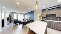 This single-floor residence showcases a thoughtfully designed open-concept layout that seamlessly connects the kitchen, dining, and living areas. The kitchen features warm wood-toned cabinetry, light countertops, and a large white island with bar seating, illuminated by striking teardrop-shaped pendant lights that serve as a focal point. The dining area sits just beyond the island, offering a round table surrounded by mid-century modern chairs, creating an inviting space for gathering. A bold navy accent wall anchors the transition between the dining and living spaces, adding depth and character to the interior. The living area is generously sized, furnished with comfortable seating and anchored by a ceiling fan. Large floor-to-ceiling windows along the far wall flood the space with natural light and provide views of the surrounding area. Light wood-toned flooring runs throughout, complementing the neutral white ceiling fitted with recessed lighting. A walk-in closet or pantry is visible through a hallway, adding practical storage to the well-appointed layout.