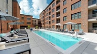 Located at 601 Meeting Street in Charleston, South Carolina, this amenity space offers a well-appointed outdoor retreat defined by a blend of classic brick architecture and contemporary design. At the heart of the courtyard sits a resort-style pool lined with lounge chairs and umbrella-shaded seating, framed on all sides by the surrounding multi-story brick and stucco building façade that creates a sense of enclosure and privacy. The pool deck is finished in large-format concrete pavers, providing a clean and durable surface ideal for year-round enjoyment.

Adjacent to the pool, a spacious open-air terrace offers an expansive gathering area with a long communal dining table and bench seating, positioned against a backdrop of exposed brick columns. This area transitions seamlessly into a covered outdoor lounge, where a wall-mounted flat-screen TV, ceiling fan, and comfortable sectional furniture create a relaxed, living-room-like atmosphere. Horizontal shiplap-style wall paneling in muted grey tones adds a refined coastal touch to the covered area.

The courtyard entry is marked by floor-to-ceiling glass doors that offer a preview of the interior lounge beyond, combining transparency and accessibility with a polished, modern aesthetic. Together, these amenity spaces provide a cohesive and functional outdoor environment suited for both social gatherings and quiet relaxation.