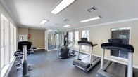 This fitness center offers a practical and well-organized workout environment designed to support a variety of exercise routines. The space is outfitted with multiple pieces of cardio equipment, including treadmills and an elliptical trainer, positioned to take advantage of the natural light streaming in through the windows. A dedicated strength training area features a multi-function cable machine, complemented by floor-to-ceiling mirrors that visually expand the room and allow users to monitor their form during workouts. The flooring throughout is a durable, dark rubberized material well-suited for high-traffic fitness use. Overhead, recessed panel lighting ensures the space is evenly and brightly illuminated. Crown molding along the ceiling adds a subtle architectural refinement to an otherwise functional space. The neutral beige walls contribute to a clean, focused atmosphere. Natural light filters in through multiple windows, providing views of the surrounding exterior and enhancing the overall sense of openness. The layout is efficient, offering enough room to move freely between equipment. The facility is contained on a single floor, making it easily accessible and straightforward to navigate.