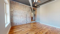 This single-floor unit in Leeds, Alabama, masterfully blends historic industrial character with modern finishes. The entry hall immediately establishes the aesthetic with a full-height exposed brick wall running alongside warm hardwood floors, while overhead ductwork and corrugated metal ceiling panels reinforce the building's industrial origins. A ceiling fan and recessed lighting provide both function and ambiance throughout the open living areas.

The living space is generously proportioned, anchored by a striking original brick column and accented by an arched brick doorway that frames transitions between rooms. Large windows allow abundant natural light to pour in, offering views of the surrounding streetscape. The kitchen features contemporary cabinetry, stainless steel and black appliances including a refrigerator, range, and microwave, with hardwood flooring continuing seamlessly from the main living area.

Two well-sized bedrooms each feature multiple windows, hardwood floors, and distinctive ceiling treatments ranging from corrugated metal to warm wood paneling. Both bathrooms are finished with light wood vanities, white tile surrounds, tub-shower combinations, and neutral tile flooring, providing a clean contrast to the raw industrial elements found throughout the rest of the space.