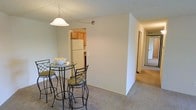 This single-floor unit offers a functional and straightforward layout designed for comfortable everyday living. The dining area is finished with neutral wall-to-wall carpeting and features a bistro-style glass-top table with wrought iron chairs positioned beneath a classic pendant light fixture, adding a subtle decorative touch to the space. A partial wall separates the dining area from the kitchen, which is visible through an open pass-through layout. The kitchen is equipped with warm wood-tone cabinetry, a white refrigerator, and light-toned countertops, providing a practical cooking space with a cozy feel. The neutral color palette throughout — featuring soft white and beige tones on the walls and ceiling — creates a clean, understated ambiance. A hallway extends from the main living area, hinting at additional rooms beyond, including what appears to be a bedroom or secondary living space. The overall design is simple and utilitarian, with an open flow between the main living, dining, and kitchen zones, making the space feel connected and easy to navigate.