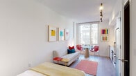 This single-floor studio is thoughtfully designed with a clean, modern aesthetic characterized by crisp white walls, light hardwood flooring, and an open layout that maximizes the sense of space. The main living area seamlessly integrates the sleeping, lounging, and dining functions. A full-sized bed anchors one end of the room, dressed in yellow and teal textiles and accompanied by closet storage along the adjacent wall. A compact yet comfortable sofa sits mid-room, paired with a small side table, while a round dining table with vibrant red lattice chairs occupies the windowed end of the space.

A large floor-to-ceiling window at the far end floods the room with natural light and provides views of the surrounding cityscape. Colorful framed artwork in gold-toned frames punctuates the white walls, and an architectural pendant light fixture adds a sculptural focal point above.

The kitchen is sleek and functional, outfitted with flat-front white cabinetry, white quartz countertops, a textured tile backsplash, and stainless steel appliances including an oven, dishwasher, and refrigerator. The bathroom features a glass-enclosed walk-in shower with floor-to-ceiling white subway tile and built-in niche shelving, finished with polished chrome hardware. The overall ambiance is light, airy, and energized by carefully chosen pops of color throughout.