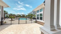 This single-story amenity building serves as the social hub of a resort-style residential community, thoughtfully designed to blend indoor comfort with outdoor living. The exterior showcases a classic coastal aesthetic, combining gray lap siding, white trim, and stacked stone accents at the column bases and fireplace surround, all contributing to a polished, cohesive architectural character.

A generously sized covered porch serves as the centerpiece of the outdoor experience, featuring exposed wood beam ceilings and large white columns resting on stone-clad bases. The porch is furnished with wicker seating and vivid blue cushions, creating an inviting lounge atmosphere with open views of the surrounding landscape and palm trees. A prominent stone fireplace anchors one end of the porch, adding warmth and a focal point for gathering.

Directly adjacent to the covered porch, a spacious paver-laid pool deck extends toward a resort-style swimming pool enclosed by sleek black iron fencing. The pool area is framed by palm trees and lounge seating, with multi-story residential buildings visible in the background, reinforcing the community setting. Large windows along the building façade allow natural light to flow into the interior spaces, which include access to a dedicated fitness center. The combination of thoughtful design details, durable materials, and resort-quality amenities makes this space a standout communal asset.