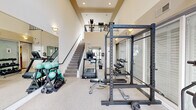 This fitness center is a thoughtfully designed, two-level workout facility that offers a comprehensive range of equipment for both cardio and strength training. The space is defined by its soaring double-height ceilings, which lend an open, airy atmosphere, and large floor-to-ceiling glass doors that connect directly to an outdoor pool area, filling the interior with natural light and providing convenient access to the amenity spaces beyond.

The main floor houses a robust selection of strength training equipment, including a full power rack with a weight bench and barbell, a multi-tiered dumbbell rack stocked with a wide range of weights, medicine balls, and kettlebells. A treadmill and additional cardio machines are centrally positioned, while full-length wall mirrors visually expand the space and support functional training. Soft carpeting covers the main workout area, and wall sconces complement the recessed lighting throughout.

A staircase leads to the upper mezzanine level, where additional cardio equipment is available, including Peloton spin bikes, an elliptical trainer, and a cable pulley machine. A wall-mounted television on the upper level adds an entertainment element for users. The overall ambiance is clean, bright, and purposefully functional, making this fitness center a well-rounded amenity suitable for a wide variety of workout routines.