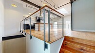 Located at 340 Rutgers St in Rochester's vibrant 14607 neighborhood, this two-story, 2-bedroom, 2-bathroom residence blends industrial character with modern finishes across two well-proportioned floors. The upper level serves as the heart of the home, showcasing a dramatic open-concept living and kitchen area with soaring ceilings, exposed steel beams painted in bold navy, visible ductwork, and structural columns that nod to the building's industrial origins. A skylight floods the space with natural light, while a row of pendant lights adds warmth above the kitchen island. The kitchen features dark cabinetry, a stainless steel range hood, and a spacious island with an extended dining surface — ideal for both cooking and entertaining. A sleek cable-railing staircase with a warm wood handrail connects the floors, and a glass-block window on the landing diffuses soft light throughout. Both bedrooms are finished with warm hardwood flooring, exposed columns and ductwork, and high clerestory windows. The two bathrooms feature tile flooring with clean, minimal finishes. Throughout, light gray walls and natural wood tones create a cohesive, contemporary aesthetic that honors the space's industrial roots.