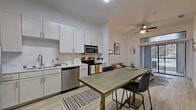 This single-floor unit is designed with a clean, contemporary aesthetic, characterized by white walls, light wood-style flooring, and a bright, open layout. The kitchen is well-equipped with stainless steel appliances, including a refrigerator, dishwasher, microwave, and range. Crisp white cabinetry extends to the ceiling, providing generous storage, while light countertops offer ample workspace. A ceiling fan with a light fixture anchors the adjoining open living and dining area, which benefits from natural light streaming through a sliding glass door that leads to an outdoor space.

The bedroom is a comfortable, well-lit room featuring a large window with blinds, a ceiling fan, and the same light wood-style flooring found throughout the unit. It includes access to a dedicated walk-in closet outfitted with wire shelving on multiple levels, offering organized storage capacity.

The bathroom is finished with large-format tile flooring, a glass-enclosed walk-in shower with subway tile surround, a single-sink vanity with chrome fixtures, and a framed mirror. The overall ambiance of the space is modern, clean, and functional, with a neutral palette that lends itself to a variety of interior styles.