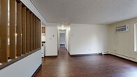 This single-floor residential unit offers a practical and functional layout designed for comfortable everyday living. Rich, dark hardwood-style flooring runs consistently throughout the space, providing a cohesive look from room to room. The open living area is generously sized, featuring a wall-mounted air conditioning unit and baseboard heating, ensuring year-round climate control. Large windows in the living and bedroom areas allow natural light to filter in while offering views of the surrounding neighborhood and mature trees.

The kitchen is positioned adjacent to the main living area, separated by a distinctive decorative partition with vertical wooden slats that adds architectural interest. It is equipped with a white gas range, a refrigerator, and warm-toned wooden cabinetry offering ample storage. A hallway connects the living space to the private areas of the unit, which include multiple bedrooms. Each bedroom features the same hardwood-style flooring, neutral walls, and baseboard heating. Several bedrooms include sliding wooden closet doors, providing practical storage solutions.

The bathroom is clean and well-appointed, featuring a full bathtub and shower combination with built-in shelving, a single-sink vanity with wooden cabinetry, and wood-look flooring. A textured ceiling is present throughout the unit, adding a classic residential character to the overall space.