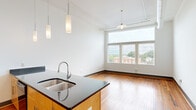 Located at 132 North Sycamore Street in Petersburg, Virginia, this two-bedroom, two-bathroom unit offers a well-designed open layout with a contemporary urban aesthetic. Rich hardwood floors run throughout the entire space, complementing the bright white walls and creating a warm, inviting ambiance.

The open-concept living and kitchen area is a standout feature, with soaring ceilings and pendant lighting that draw the eye upward. The kitchen is appointed with a large island featuring dark granite countertops, light maple cabinetry, black stainless appliances, and a double-basin sink — all seamlessly connected to the spacious living area. A large multi-pane window floods the living room with natural light and offers views of the surrounding cityscape.

A long, well-lit hallway lined with track lighting connects the main living space to the private quarters. Both bedrooms continue the hardwood flooring and feature clean white walls, ceiling fans, and ample natural light. One bedroom includes a double-door closet, while the primary bedroom benefits from an en-suite bathroom with a granite-topped vanity, maple cabinetry, and a tub-shower combination. The second bathroom mirrors this design with matching finishes. A dedicated closet provides additional storage throughout.