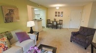 This single-floor residence features an open-concept layout that seamlessly connects the living, dining, and kitchen areas into one cohesive, functional space. The living area is carpeted and comfortably sized, providing room for a sofa arrangement and coffee table, with warm beige and yellow-green accent walls adding a welcoming touch of color.

A peninsula counter separates the kitchen from the main living space, offering a natural transition between the two areas while maintaining visual openness. The kitchen features white cabinetry, tile flooring, and standard appliances, making efficient use of its compact footprint.

Adjacent to the kitchen, the dining area accommodates a round bistro-style table with chairs, ideal for casual meals. A pendant light fixture overhead provides focused illumination, while a grouping of framed wall art adds a decorative element to the space.

The entryway, visible near the dining area, features tile flooring and a solid front door. Overall, the unit presents a clean, neutral palette with practical finishes, designed to balance comfort and functionality in a straightforward, well-organized floor plan.